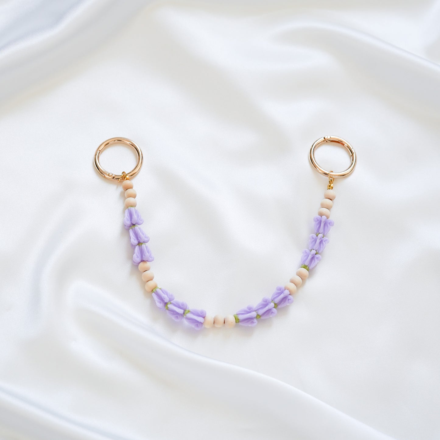 Delicate bag lei featuring puprle crown flower and tan beads in sets of three with gold rings on either end on a white fabric background