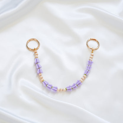 Delicate bag lei featuring puprle crown flower and tan beads in sets of three with gold rings on either end on a white fabric background