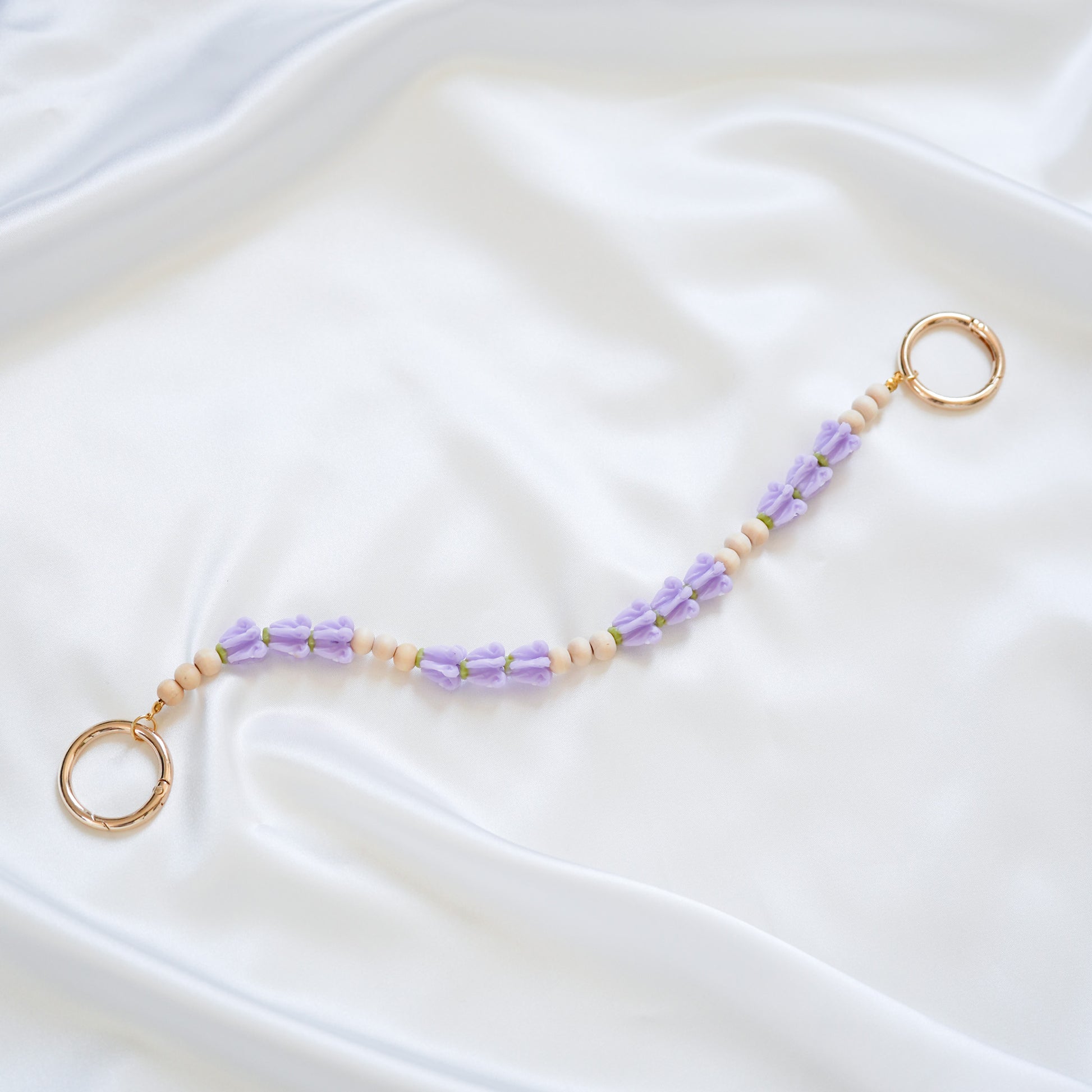 Delicate bag lei featuring puprle crown flower and tan beads in sets of three with gold rings on either end on a white fabric background