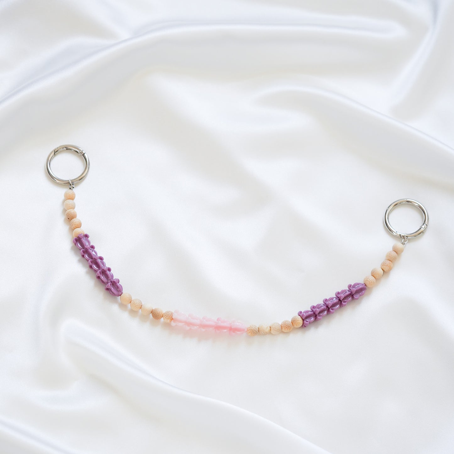 Delicate bag lei featuring purple crown flower, pink crown flower and tan beads in sets of five with gold rings on either end on a white fabric background