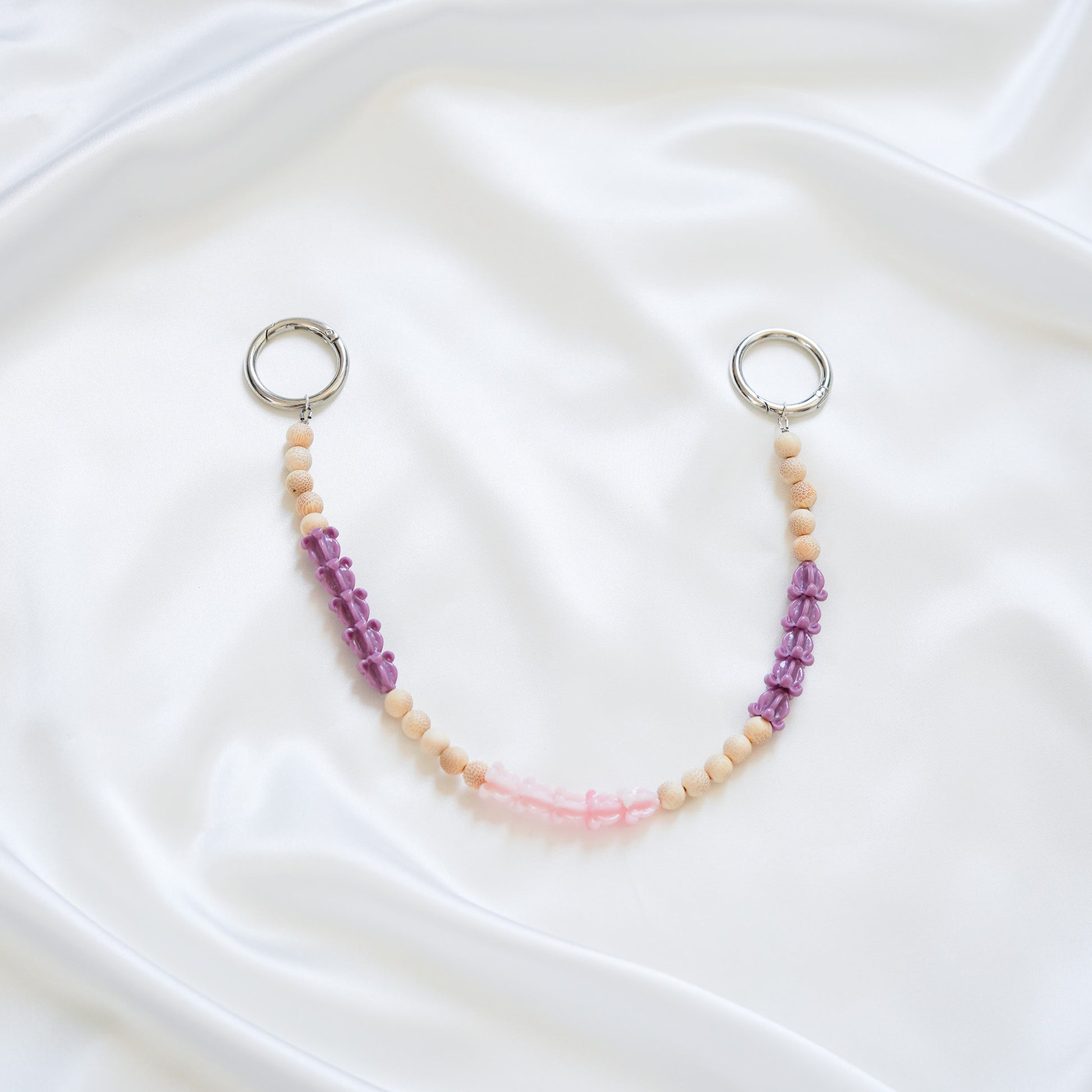 Delicate bag lei featuring purple crown flower, pink crown flower and tan beads in sets of five with gold rings on either end on a white fabric background