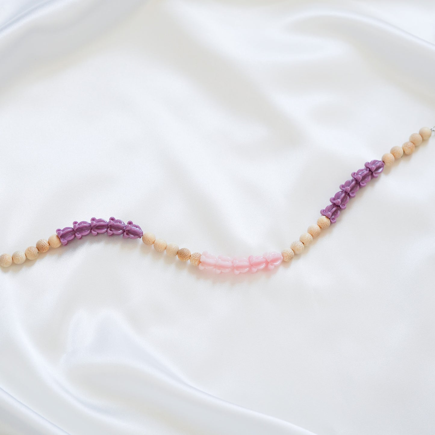 Close up of delicate bag lei featuring purple crown flower, pink crown flower and tan beads in sets of five with gold rings on either end on a white fabric background