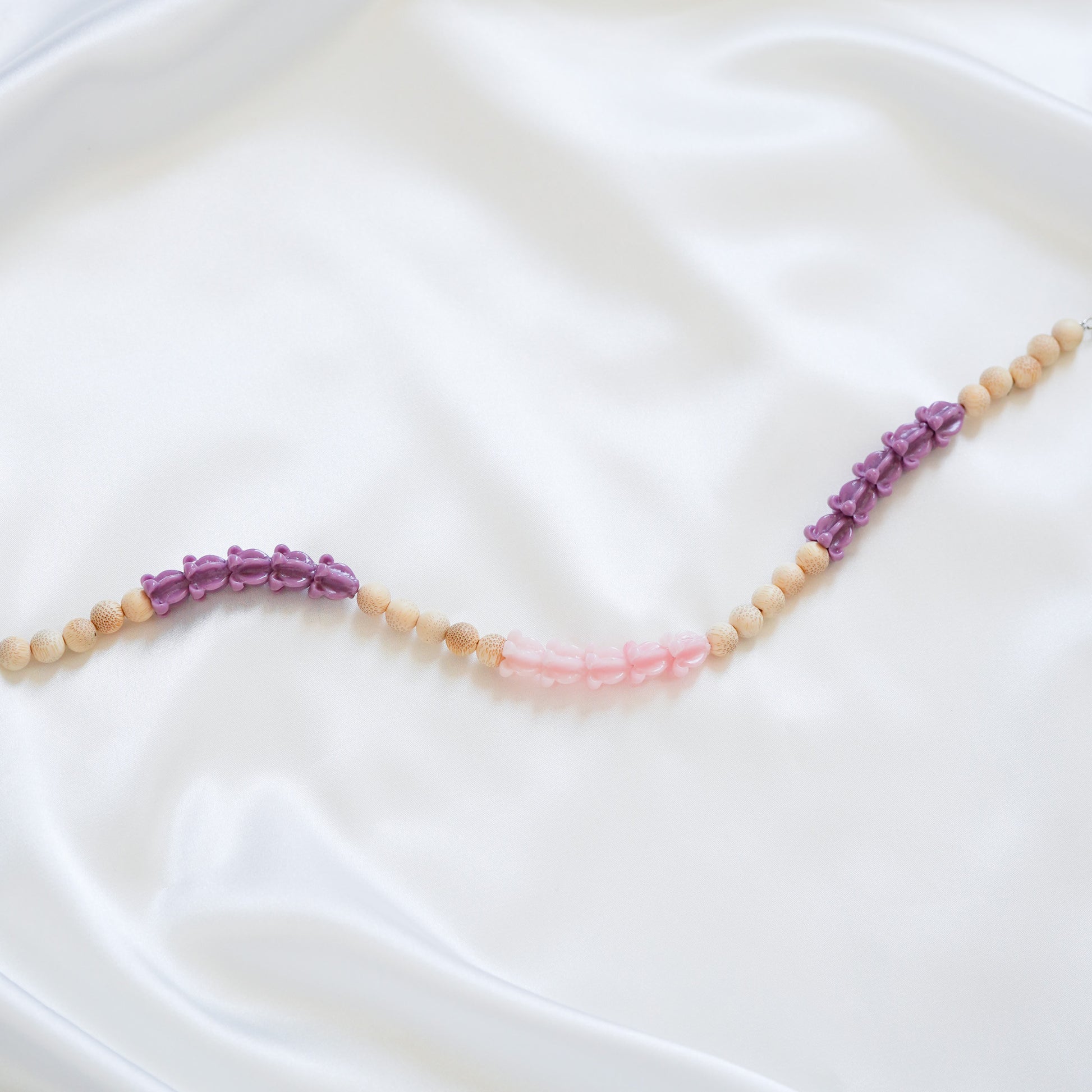 Close up of delicate bag lei featuring purple crown flower, pink crown flower and tan beads in sets of five with gold rings on either end on a white fabric background