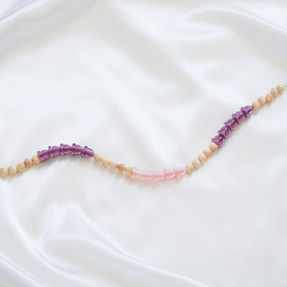 Close up of delicate bag lei featuring purple crown flower, pink crown flower and tan beads in sets of five with gold rings on either end on a white fabric background