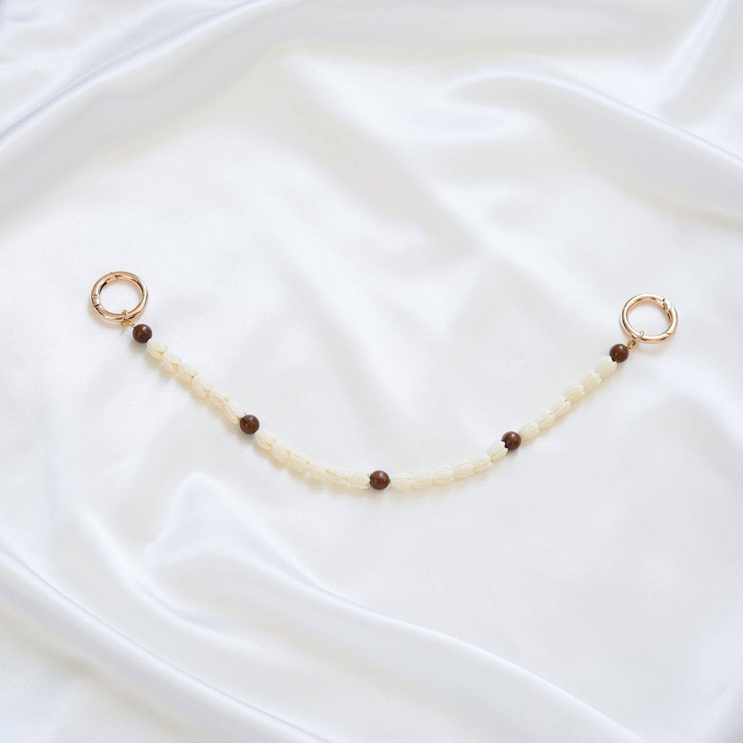 Delicate bag lei featuring white pikake flower beads and dark brown circle beads with gold rings on either end on a white fabric background