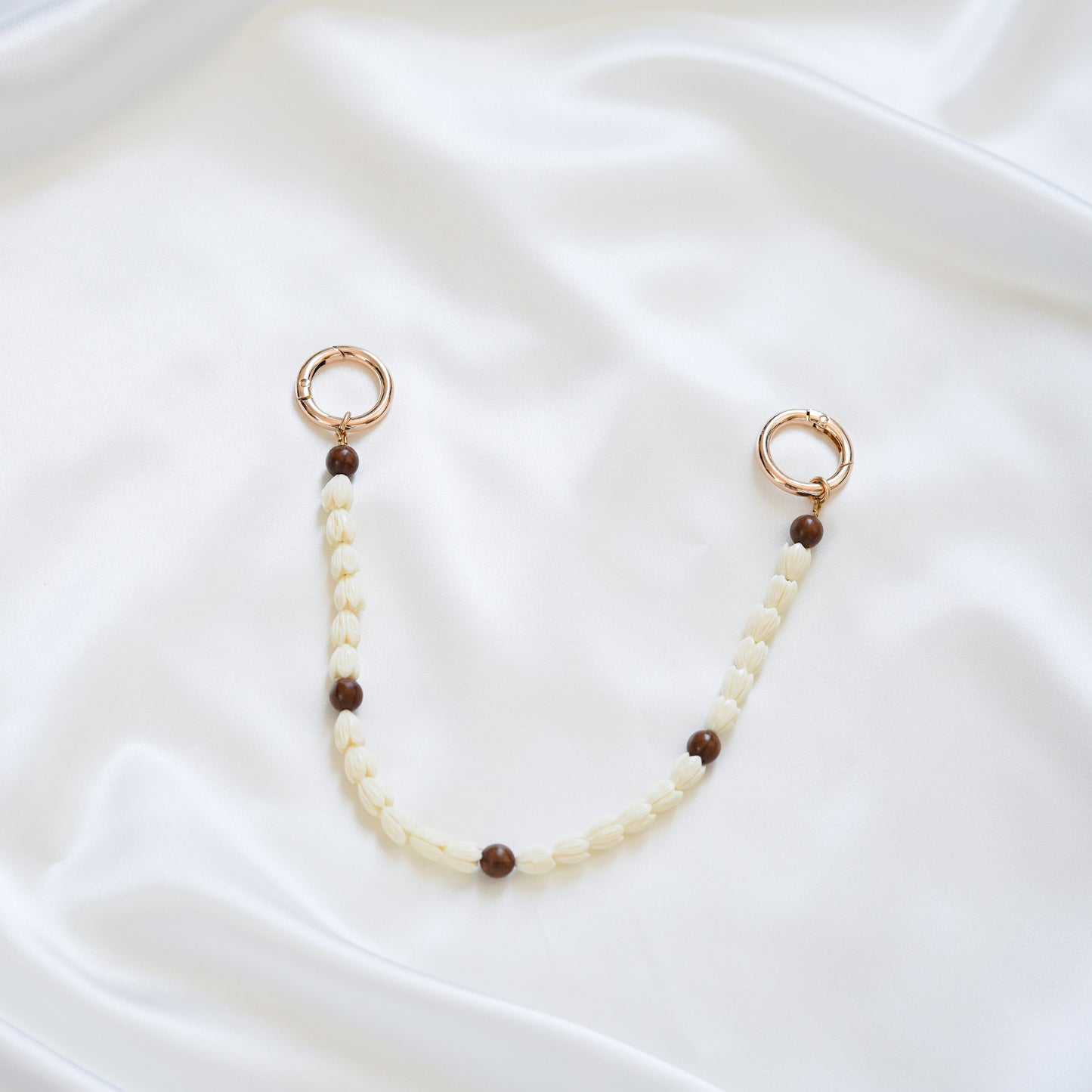Delicate bag lei featuring white pikake flower beads and dark brown circle beads with gold rings on either end on a white fabric background