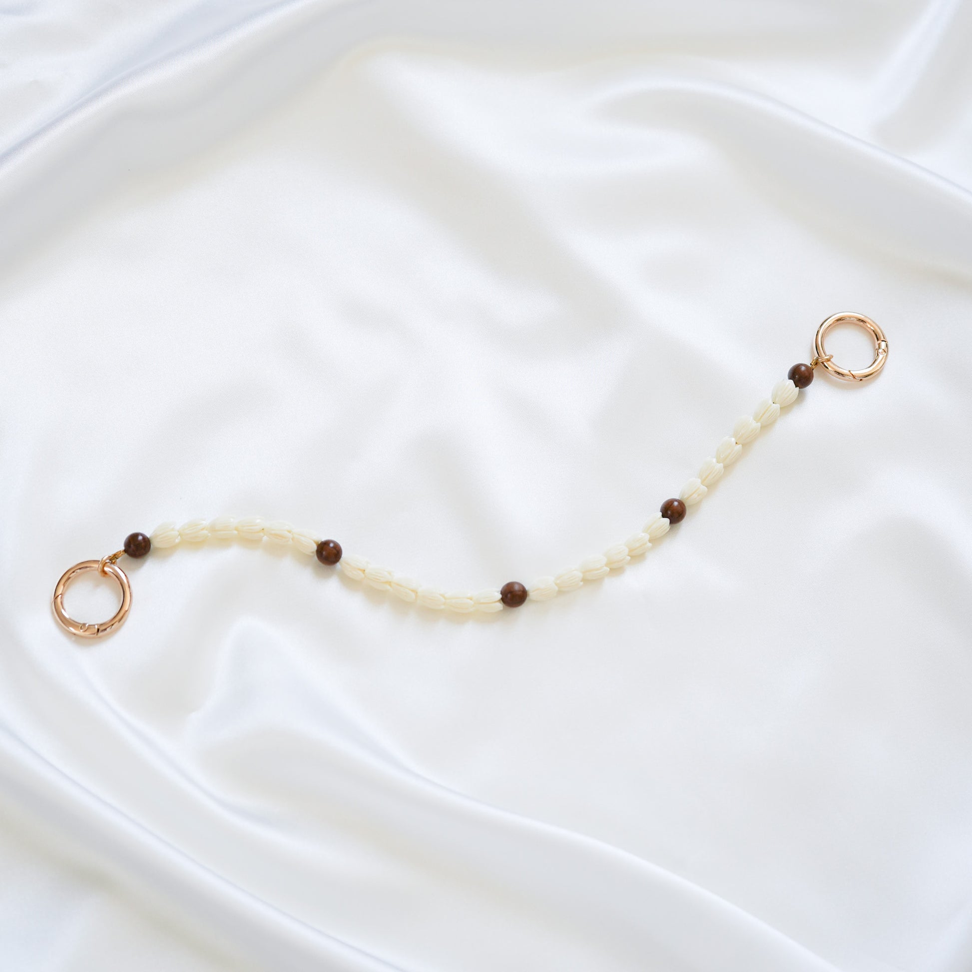 Delicate bag lei featuring white pikake flower beads and dark brown circle beads with gold rings on either end on a white fabric background