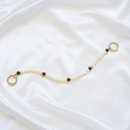 Delicate bag lei featuring white pikake flower beads and dark brown circle beads with gold rings on either end on a white fabric background
