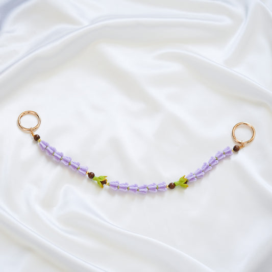 Delicate bag lei featuring purple crown flower beads and green pakalana flower beads with gold rings on either end on a white fabric background