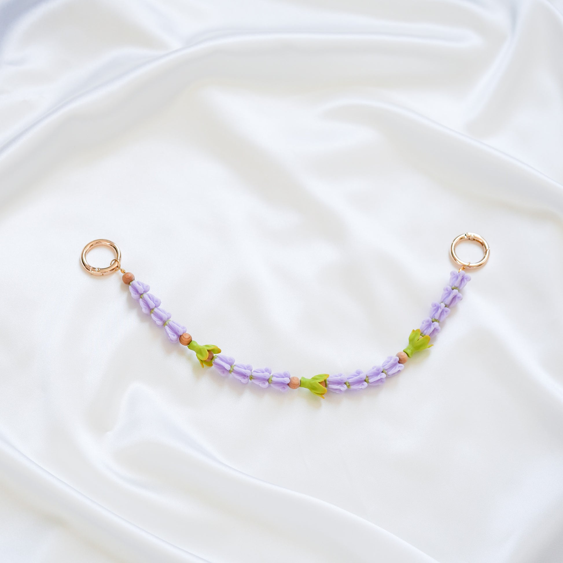 Delicate bag lei featuring purple crown flower beads and three green pakalana flower beads on a white fabric background