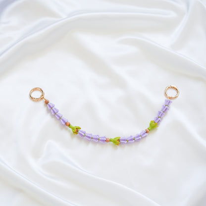 Delicate bag lei featuring purple crown flower beads and three green pakalana flower beads on a white fabric background