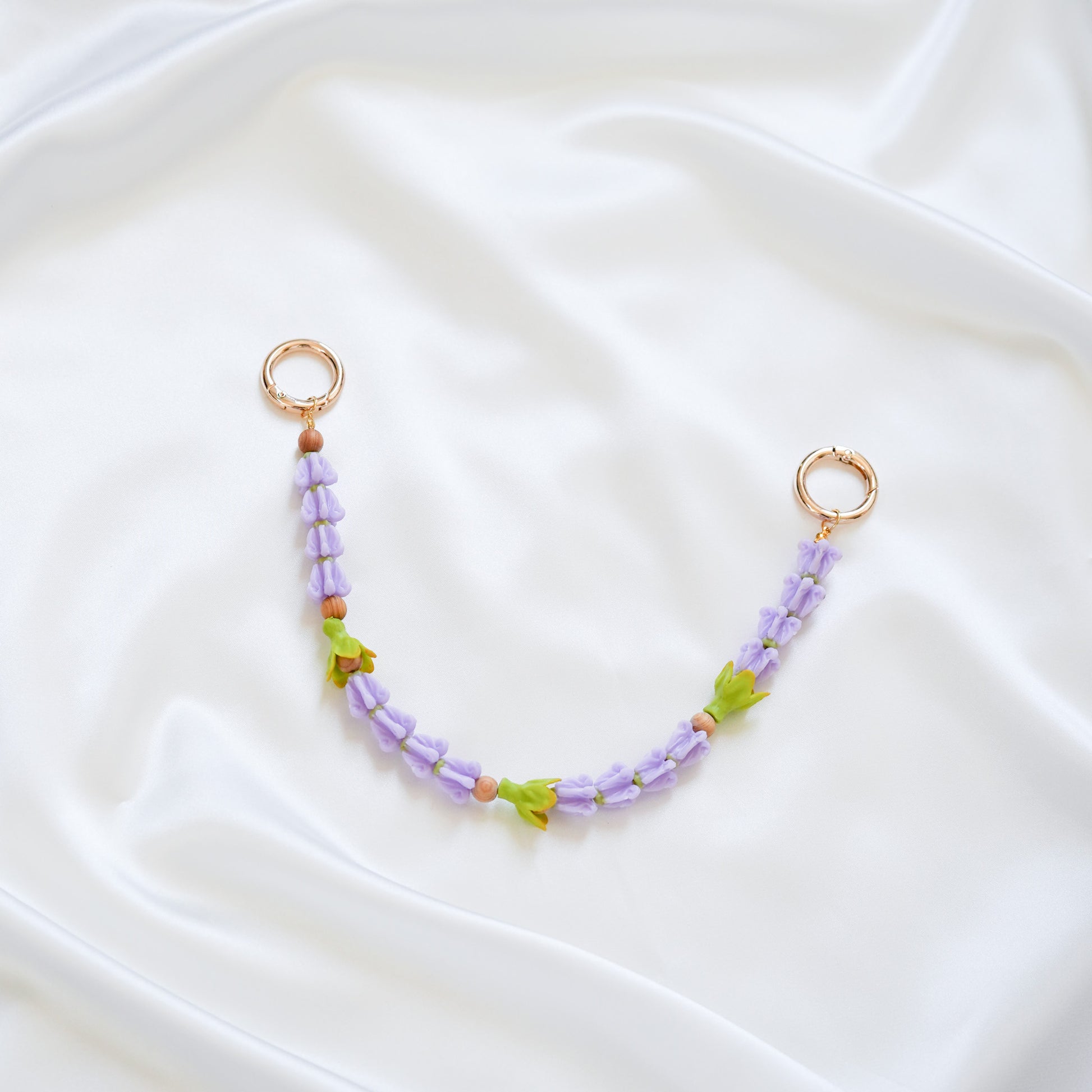 Delicate bag lei featuring purple crown flower beads and three green pakalana flower beads on a white fabric background