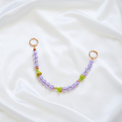 Delicate bag lei featuring purple crown flower beads and three green pakalana flower beads on a white fabric background