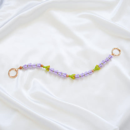 Delicate bag lei featuring purple crown flower beads and three green pakalana flower beads on a white fabric background