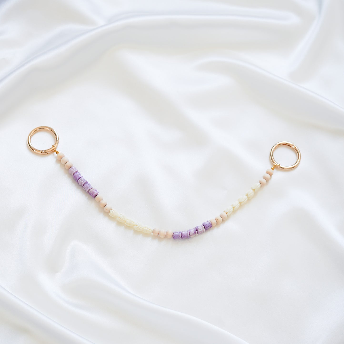 Delicate bag lei featuring small purple crown flower and white pikake flower beads with gold rings on either end on a white fabric background