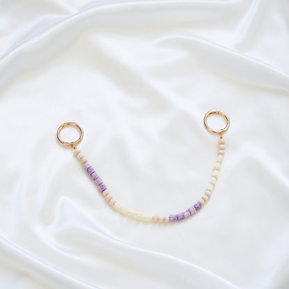 Delicate bag lei featuring small purple crown flower and white pikake flower beads with gold rings on either end on a white fabric background