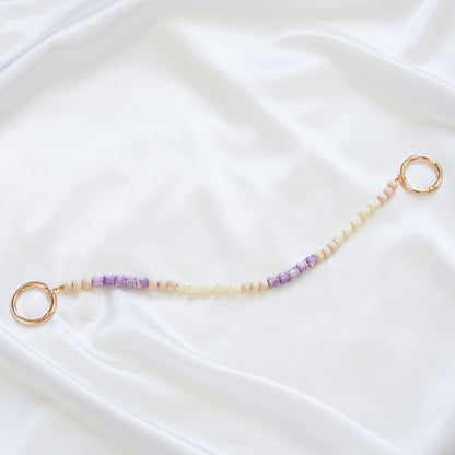 Delicate bag lei featuring small purple crown flower and white pikake flower beads with gold rings on either end on a white fabric background