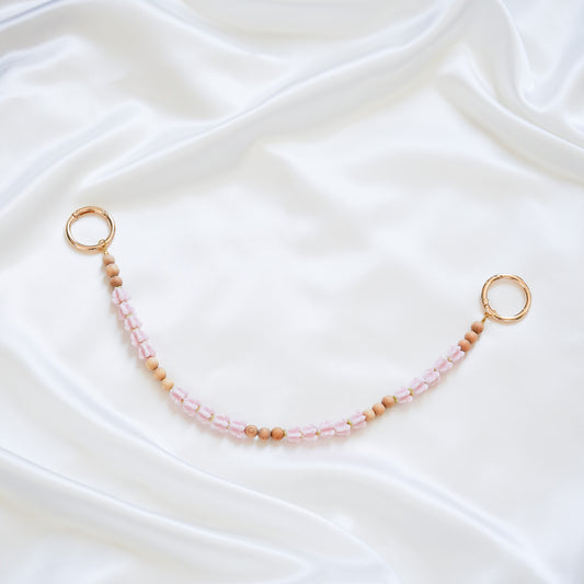 Delicate bag lei featuring pink crown flower and tan beads with gold rings on either end on a white fabric background