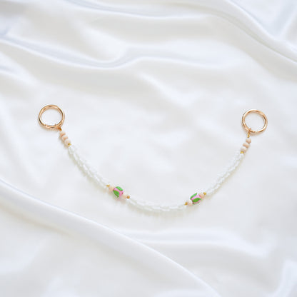 Delicate bag lei featuring white crown flower and two pink rose beads with gold rings on each end on a white fabric background