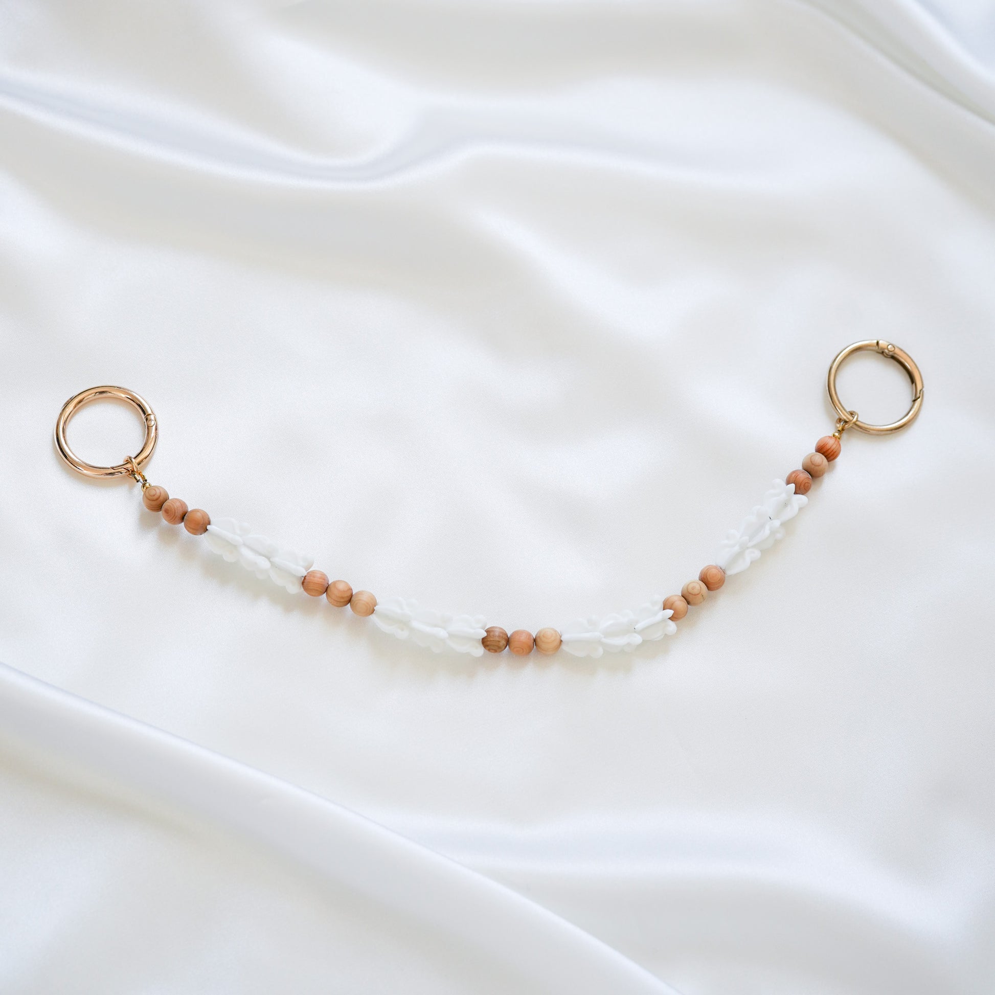 Delicate bag lei featuring white crown flower beads and tan beads alternating in sets of three with gold rings on each end on a white fabric background