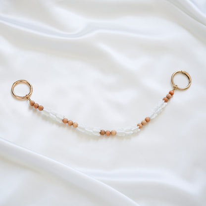 Delicate bag lei featuring white crown flower beads and tan beads alternating in sets of three with gold rings on each end on a white fabric background