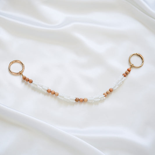 Delicate bag lei featuring white crown flower beads and tan beads alternating in sets of three with gold rings on each end on a white fabric background