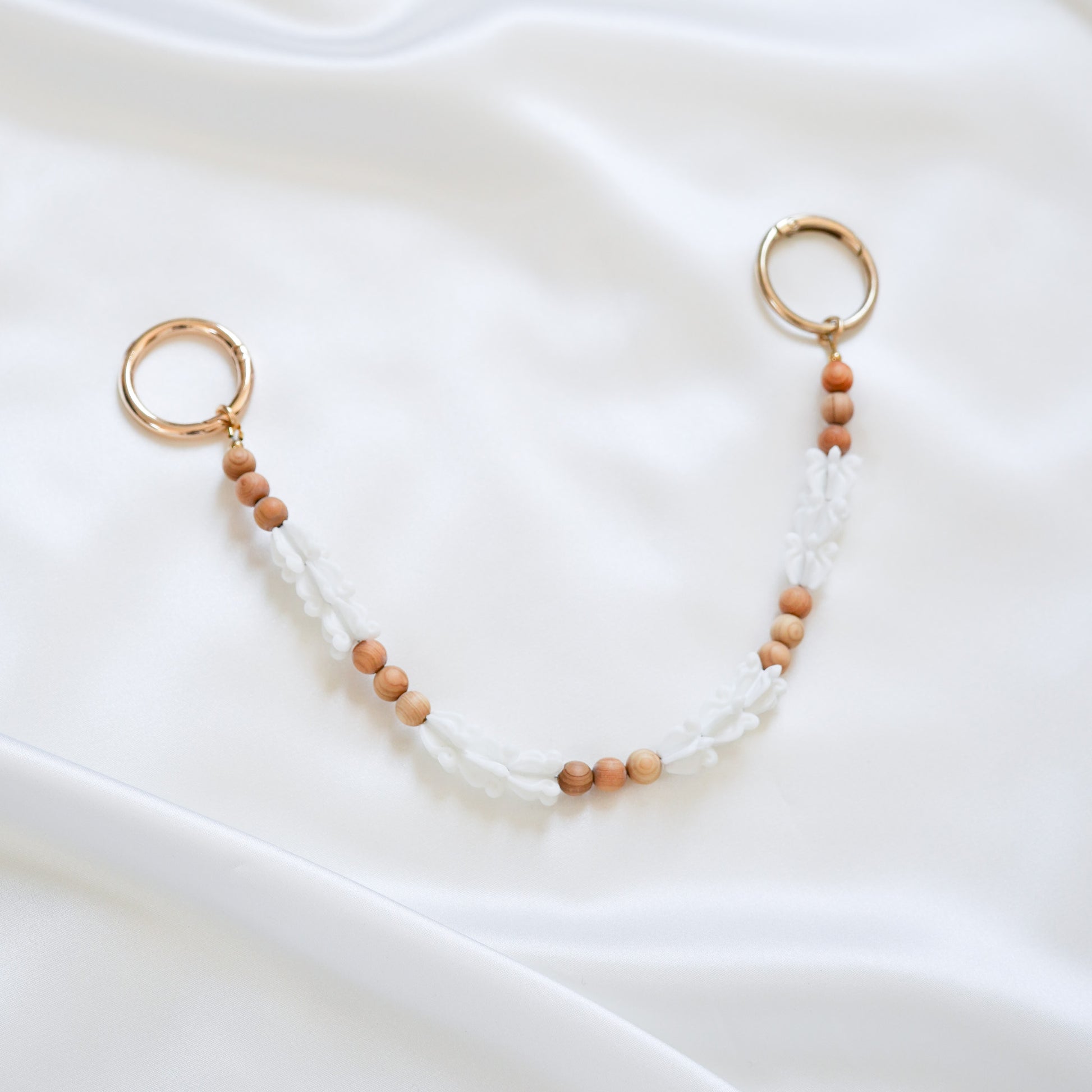 Delicate bag lei featuring white crown flower beads and tan beads alternating in sets of three with gold rings on each end on a white fabric background