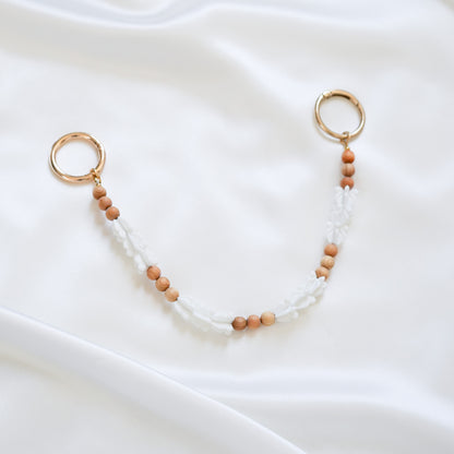 Delicate bag lei featuring white crown flower beads and tan beads alternating in sets of three with gold rings on each end on a white fabric background