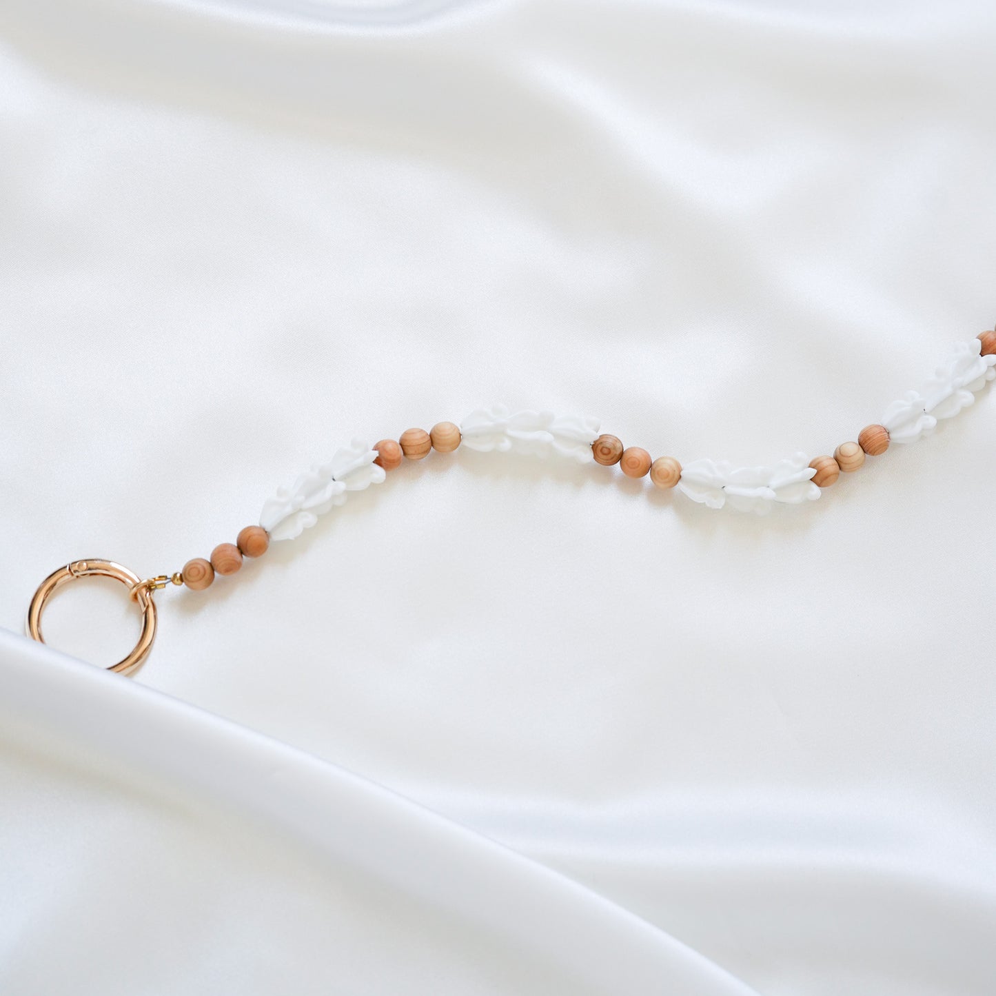 Close up of delicate bag lei featuring white crown flower beads and tan beads alternating in sets of three with gold rings on each end on a white fabric background