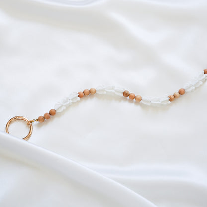 Close up of delicate bag lei featuring white crown flower beads and tan beads alternating in sets of three with gold rings on each end on a white fabric background