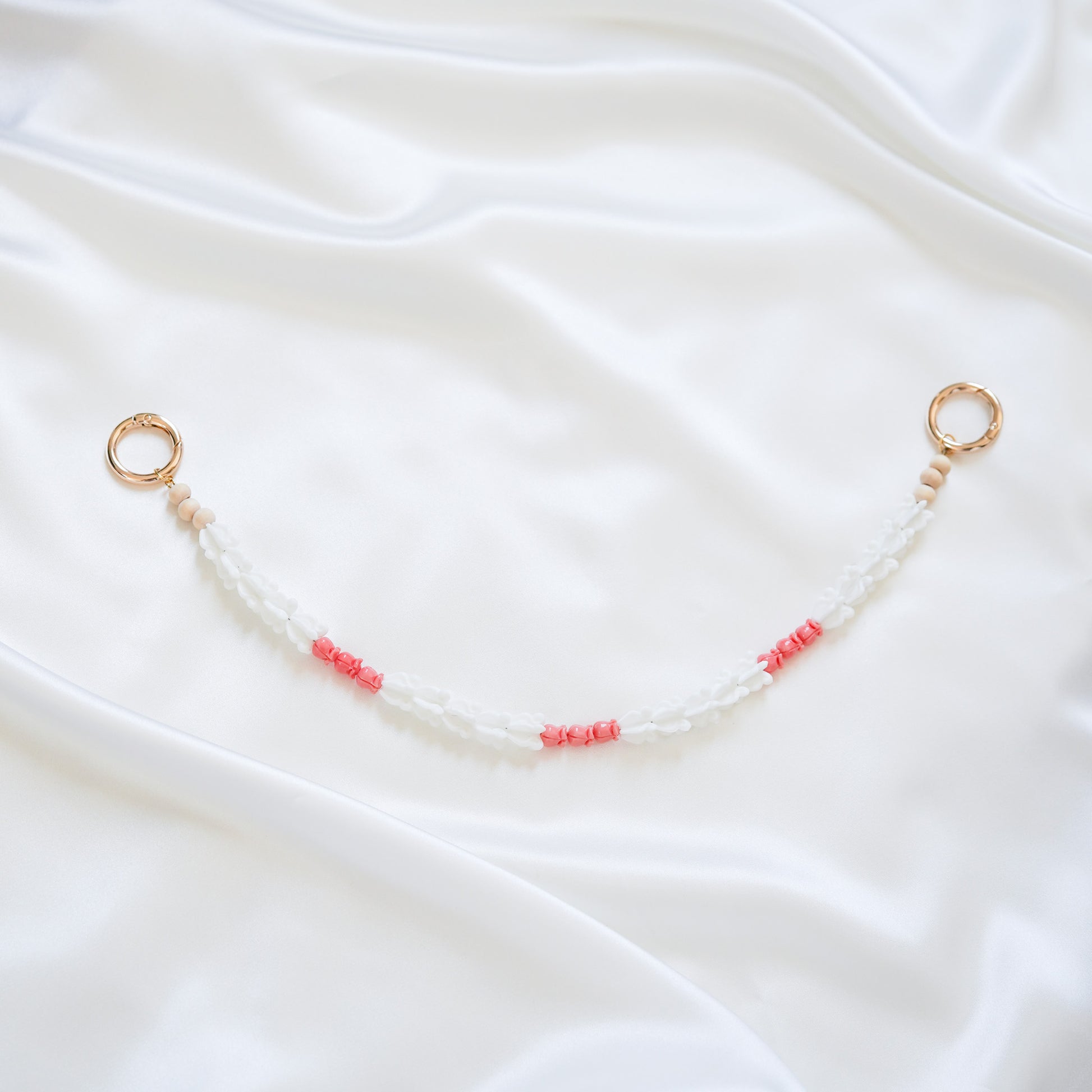 Delicate bag lei featuring white crown flower beads and small pink rose beads alternating with gold rings on each end on a white fabric background