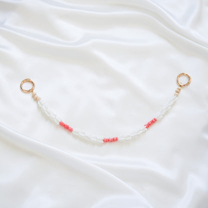 Delicate bag lei featuring white crown flower beads and small pink rose beads alternating with gold rings on each end on a white fabric background