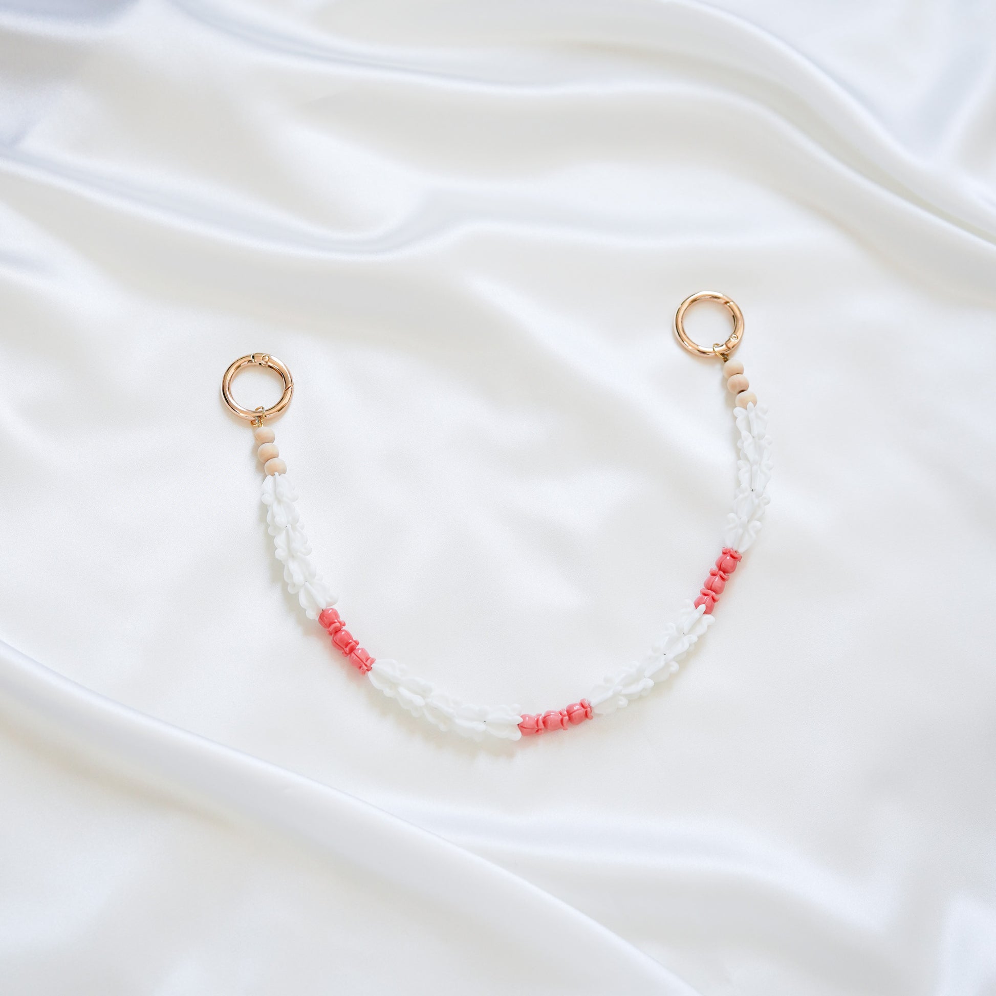 Delicate bag lei featuring white crown flower beads and small pink rose beads alternating with gold rings on each end on a white fabric background