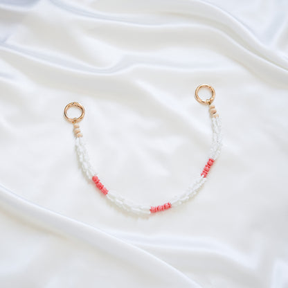 Delicate bag lei featuring white crown flower beads and small pink rose beads alternating with gold rings on each end on a white fabric background