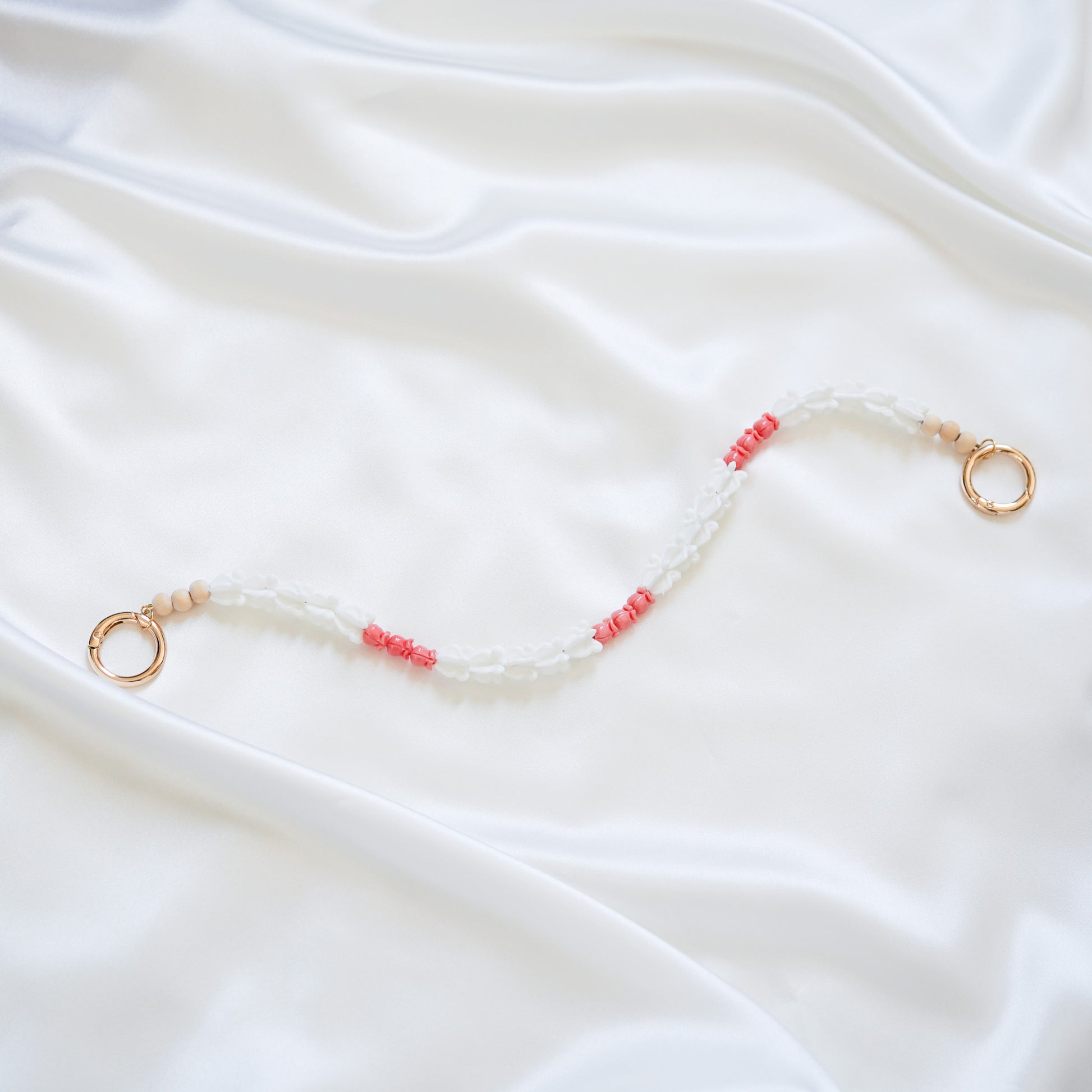 Delicate bag lei featuring white crown flower beads and small pink rose beads alternating with gold rings on each end on a white fabric background