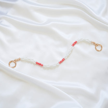 Delicate bag lei featuring white crown flower beads and small pink rose beads alternating with gold rings on each end on a white fabric background