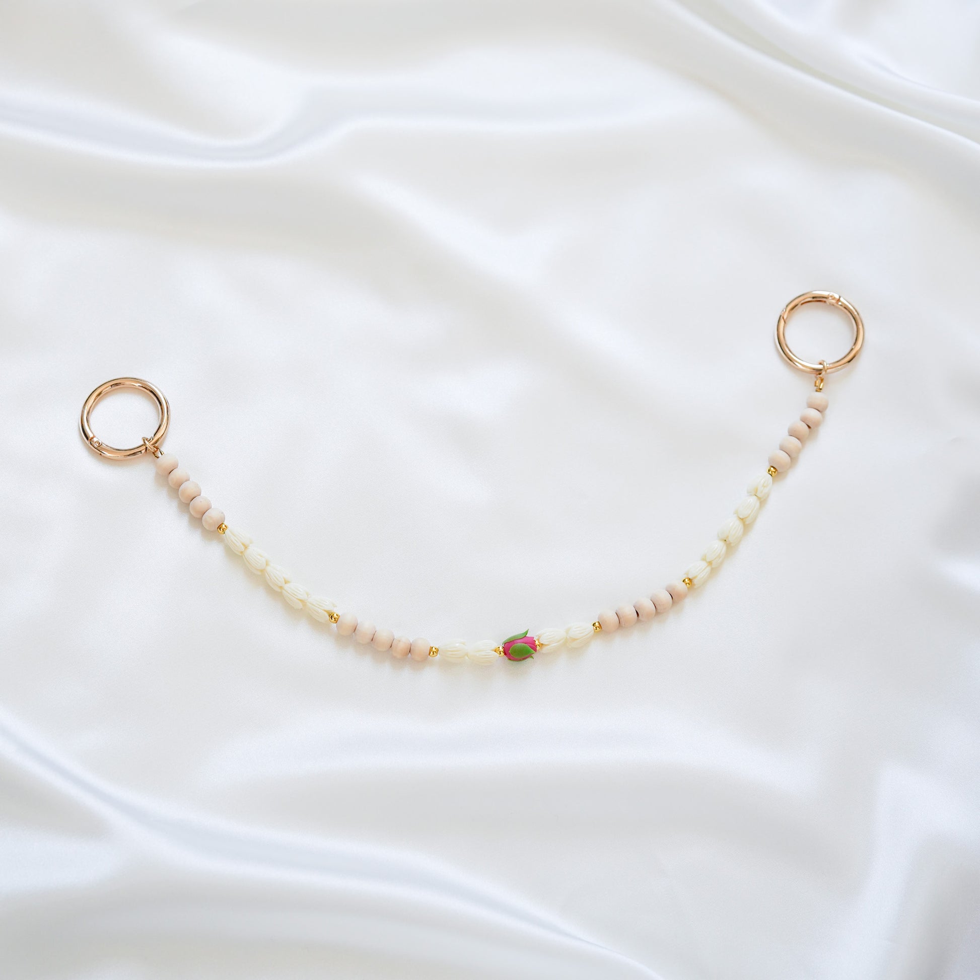 Delicate bag lei featuring white pikake flower beads and light tan beads with a pink rose bead in the middle with gold rings on each end on a white fabric background