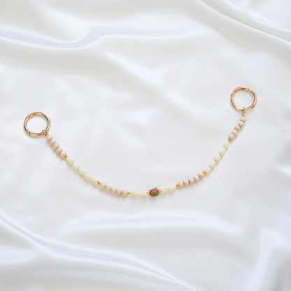 Delicate bag lei featuring white pikake flower beads and light tan beads with a pink rose bead in the middle with gold rings on each end on a white fabric background