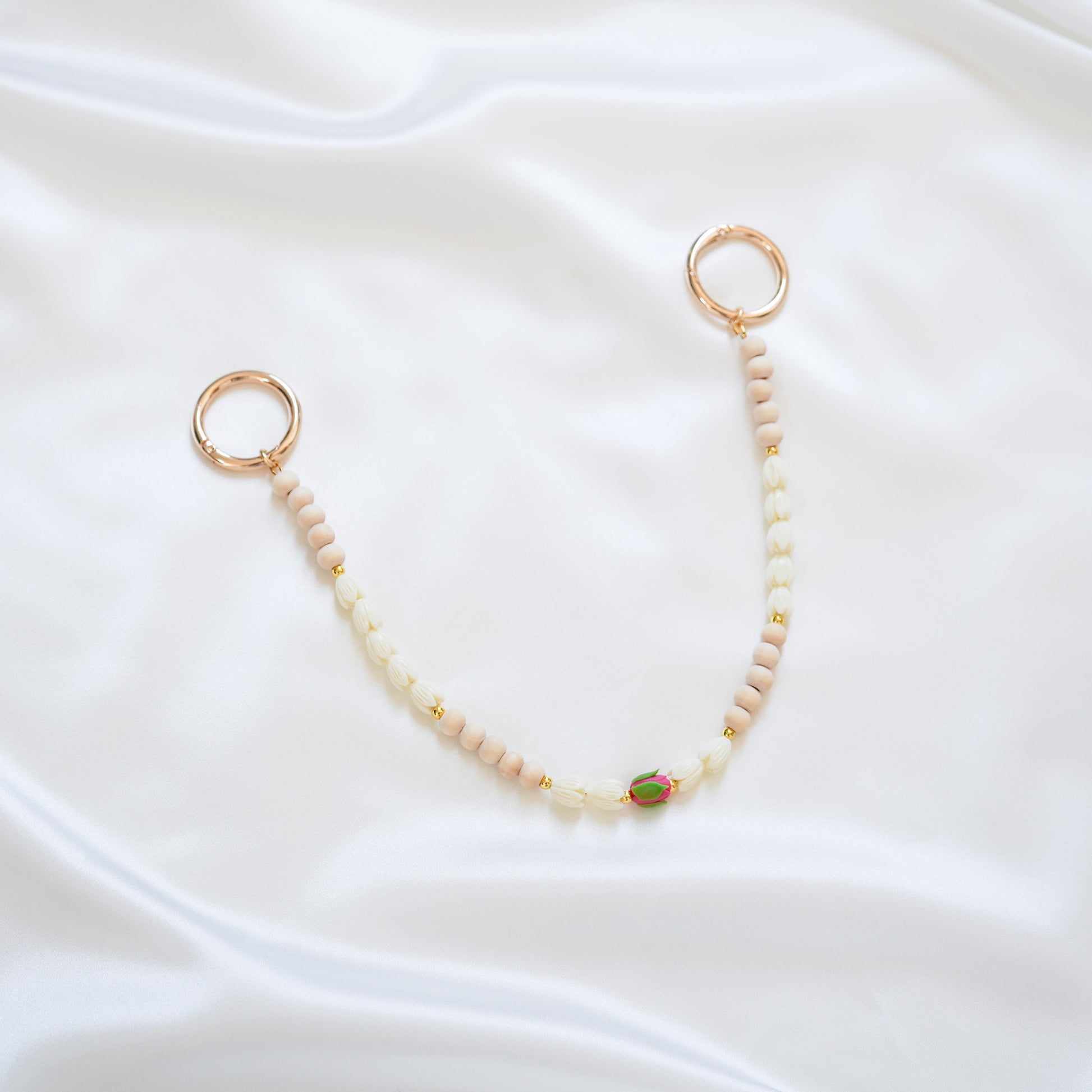 Delicate bag lei featuring white pikake flower beads and light tan beads with a pink rose bead in the middle with gold rings on each end on a white fabric background