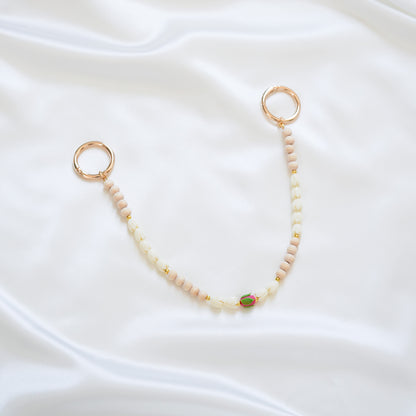 Delicate bag lei featuring white pikake flower beads and light tan beads with a pink rose bead in the middle with gold rings on each end on a white fabric background