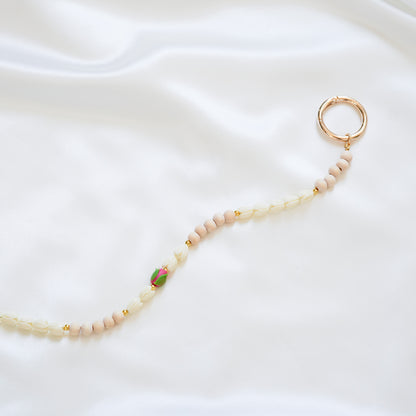 Close up of delicate bag lei featuring white pikake flower beads and light tan beads with a pink rose bead in the middle with gold rings on each end on a white fabric background