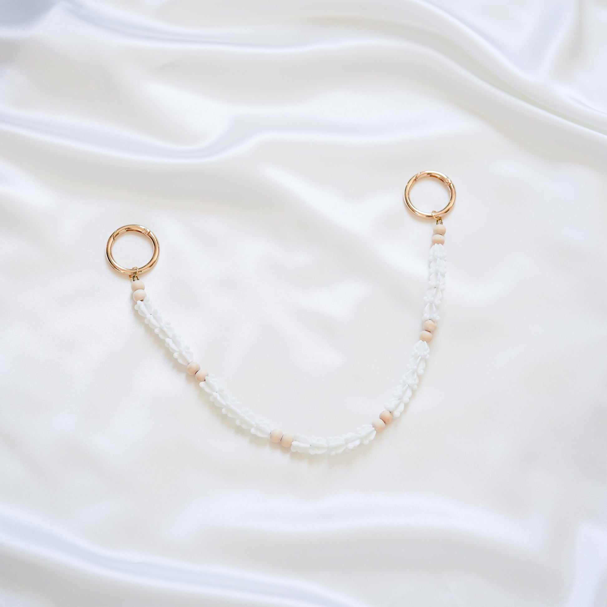 Delicate bag lei featuring white crown flower beads and light tan beads with gold rings on each end on a white fabric background