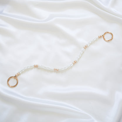 Delicate bag lei featuring white crown flower beads and light tan beads with gold rings on each end on a white fabric background