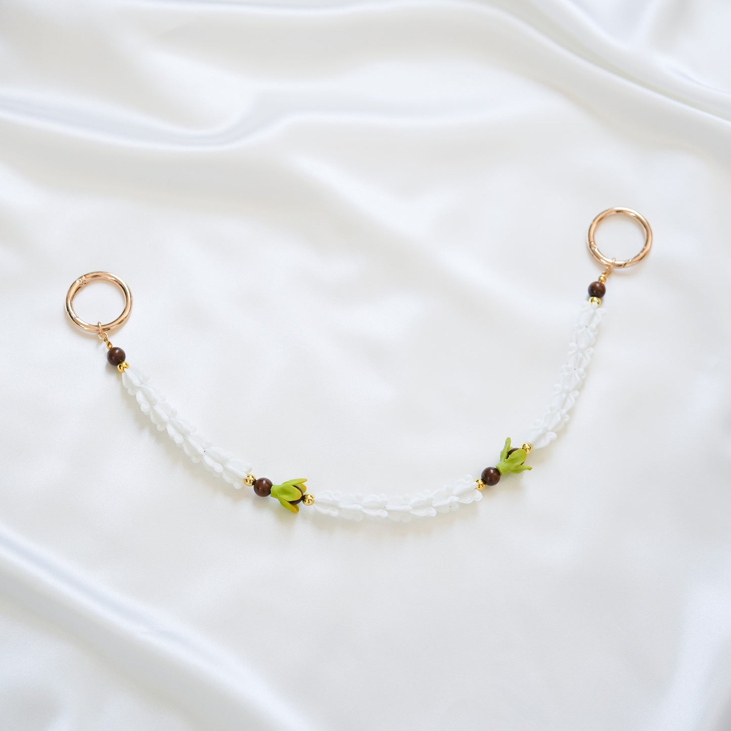 Delicate bag lei featuring white crown flower and two green pakalana flower beads and dark circle beads intertwined with gold rings on each end on a white fabric background