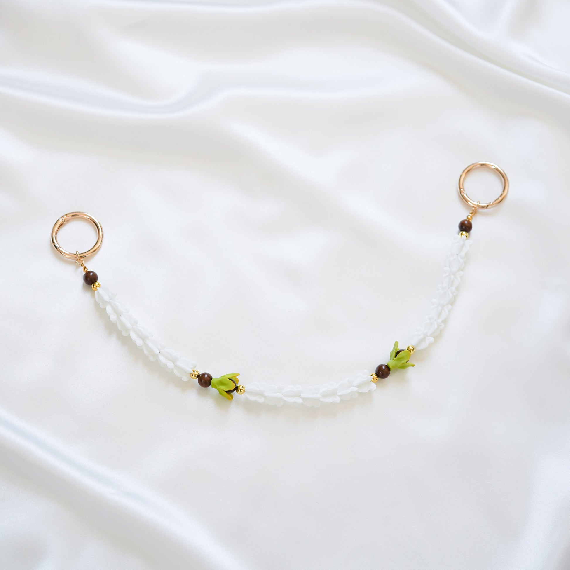 Delicate bag lei featuring white crown flower and two green pakalana flower beads and dark circle beads intertwined with gold rings on each end on a white fabric background