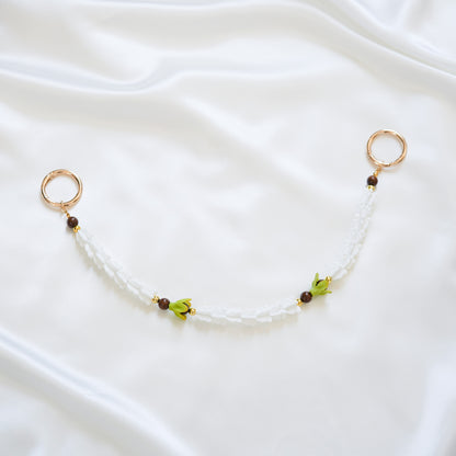 Delicate bag lei featuring white crown flower and two green pakalana flower beads and dark circle beads intertwined with gold rings on each end on a white fabric background