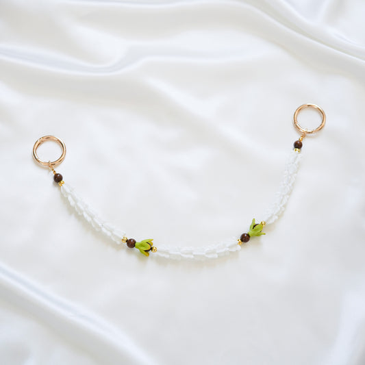 Delicate bag lei featuring white crown flower and two green pakalana flower beads and dark circle beads intertwined with gold rings on each end on a white fabric background
