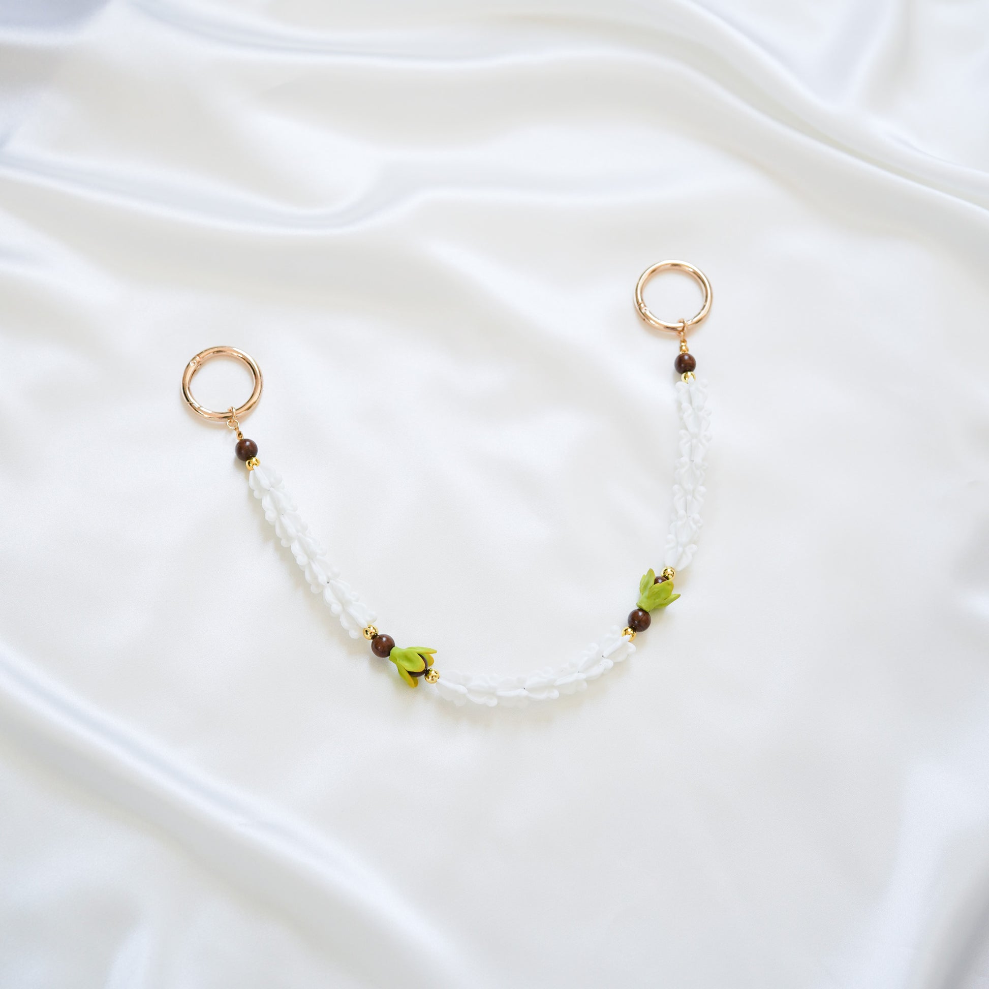 Delicate bag lei featuring white crown flower and two green pakalana flower beads and dark circle beads intertwined with gold rings on each end on a white fabric background