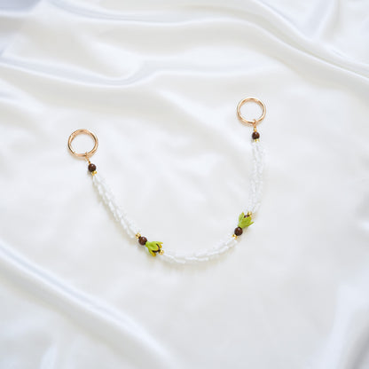 Delicate bag lei featuring white crown flower and two green pakalana flower beads and dark circle beads intertwined with gold rings on each end on a white fabric background