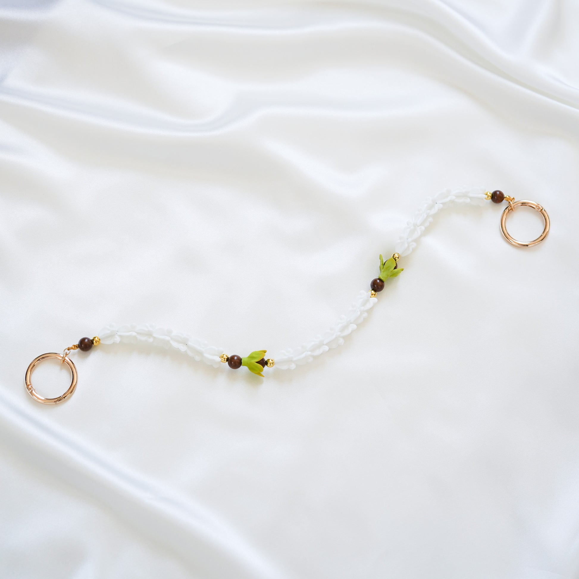 Delicate bag lei featuring white crown flower and two green pakalana flower beads and dark circle beads intertwined with gold rings on each end on a white fabric background